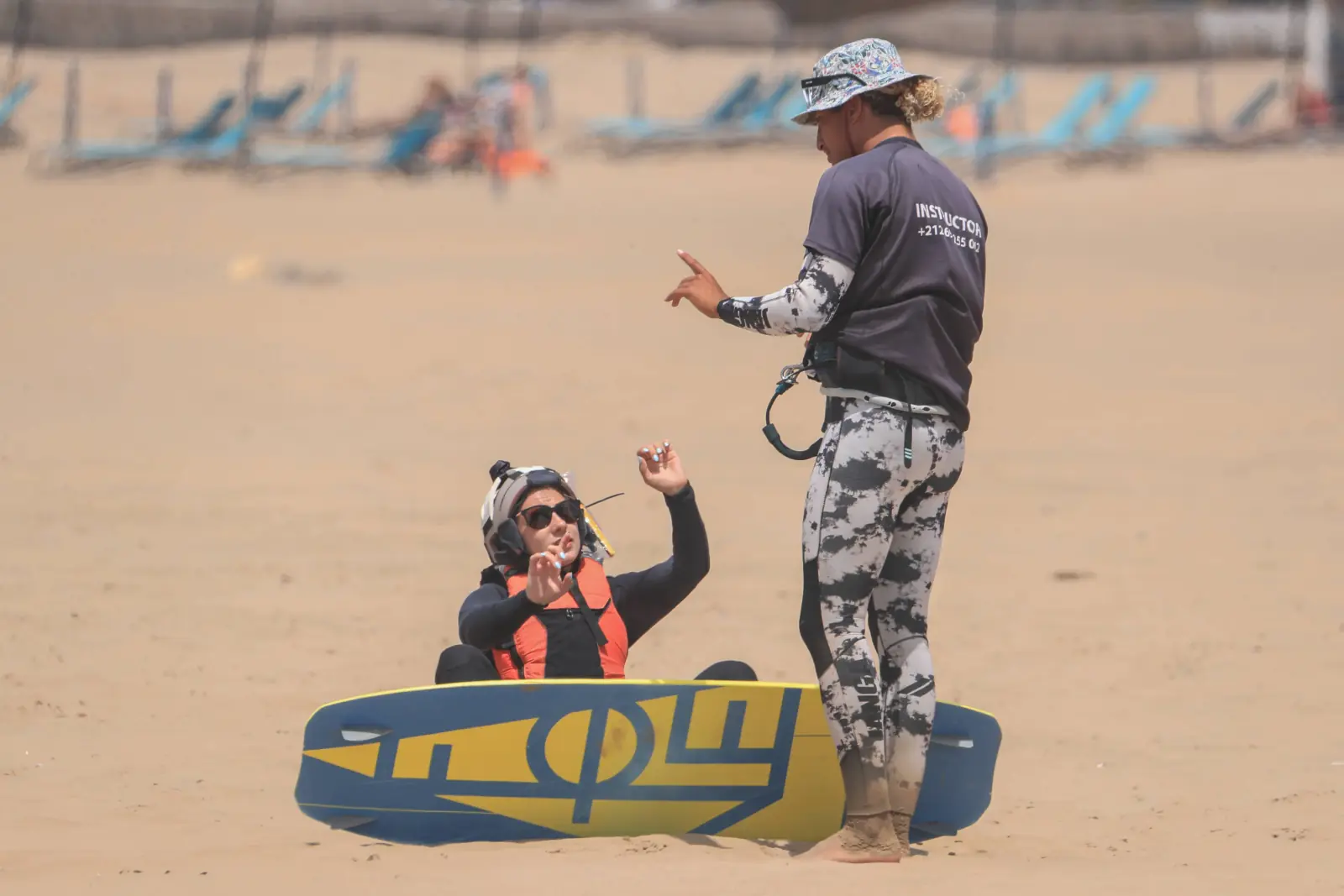 An instructor in a patterned wetsuit talks to a student sitting on the sand beside a kiteboard.