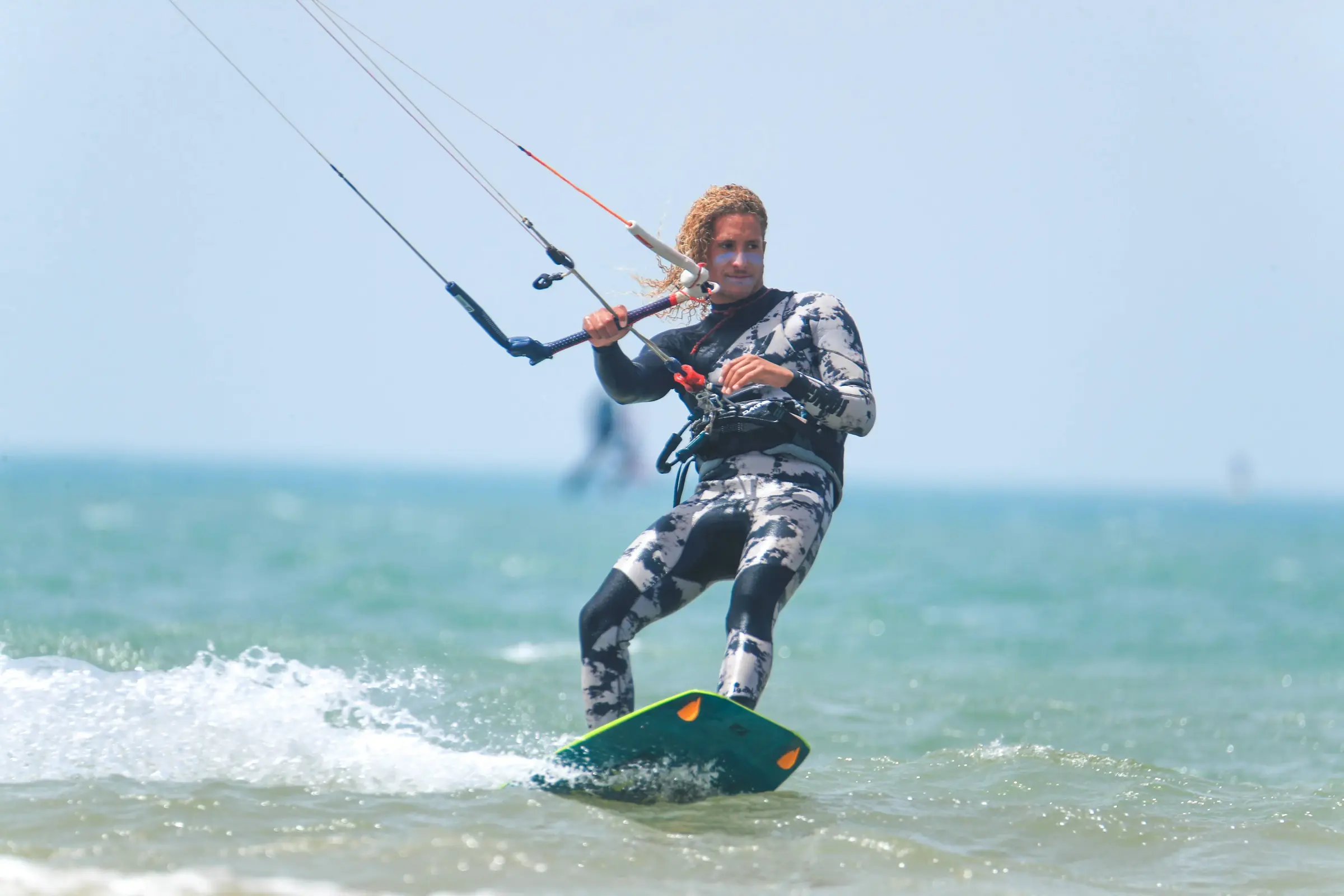 Un kitesurfeur en combinaison à motifs gris et blancs glisse sur une planche verte sur une eau turquoise, la barre de pilotage en main.