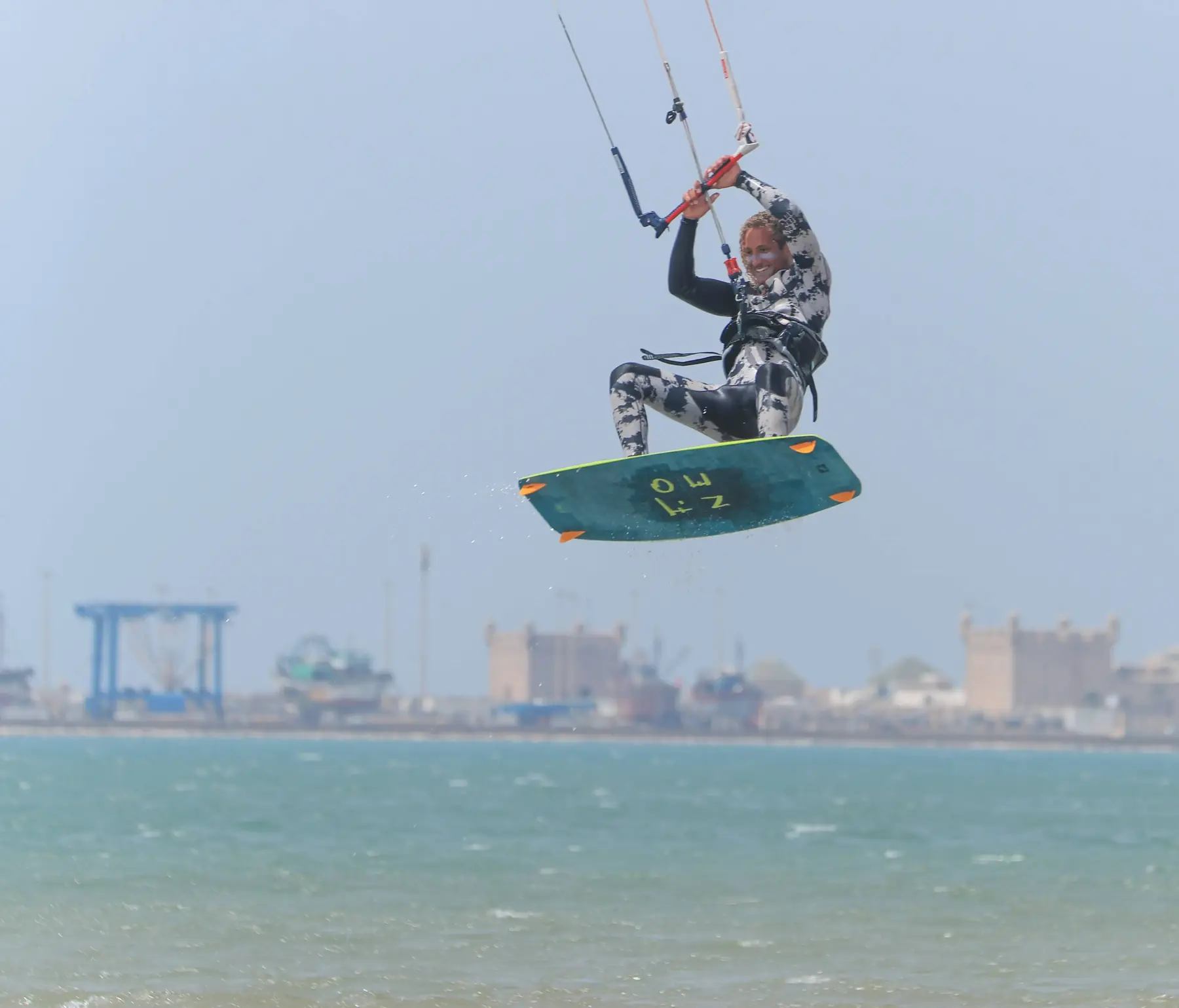 A kitesurfer in the air holding the bar above a turquoise bay, with harbour cranes and stone buildings on the shore behind.
