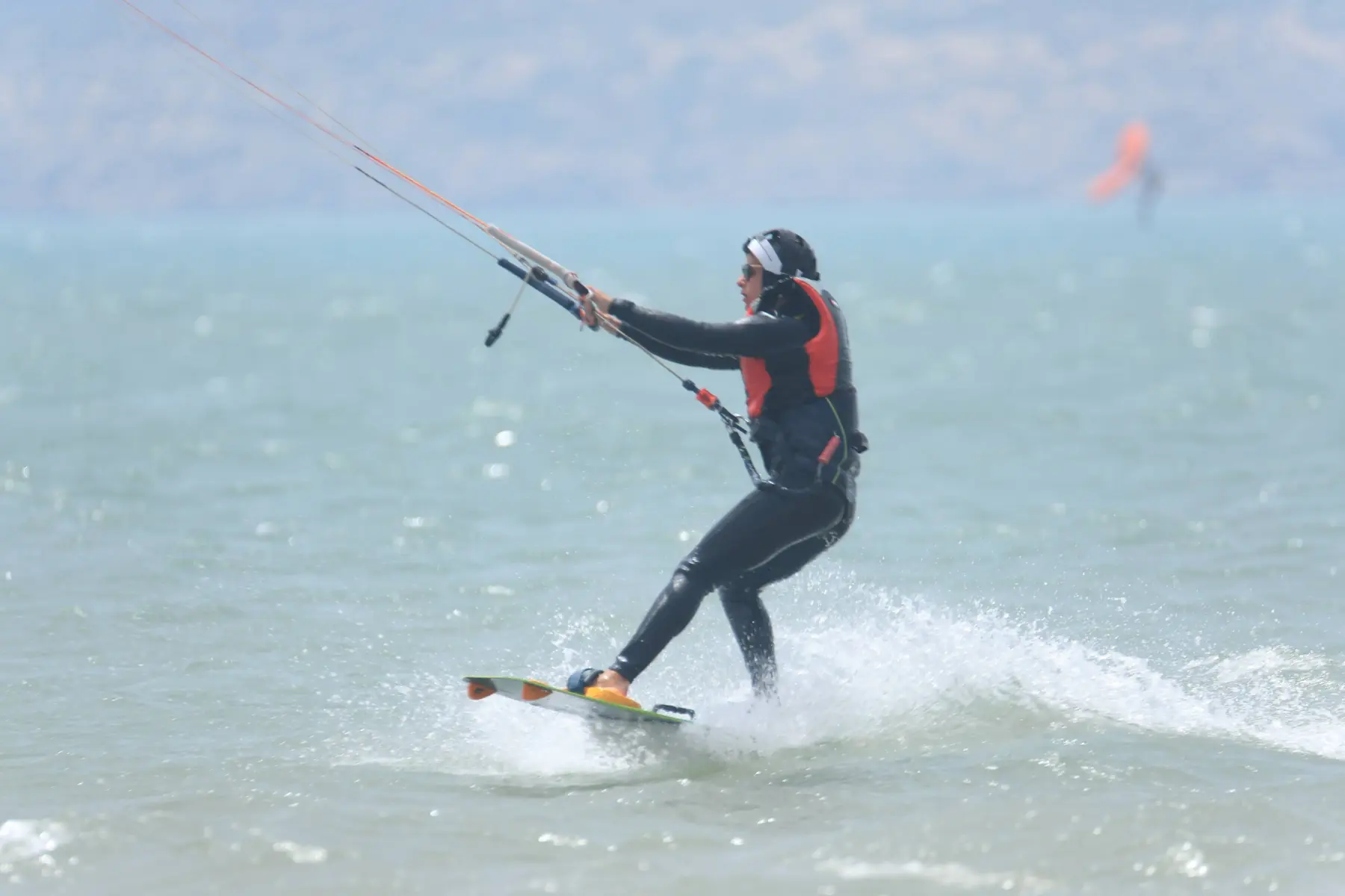 A kitesurfer in a black wetsuit and orange harness riding a board at speed, with a kite visible in the distance.